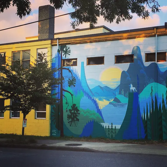 Street art mural of a mountain valley with blue colour tones and green colour tones. A wolf standing, back towards us, on one of the rock formations staring at the yellow sunset with a blue sky.